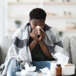 Sick black guy sitting at couch, sneezing
